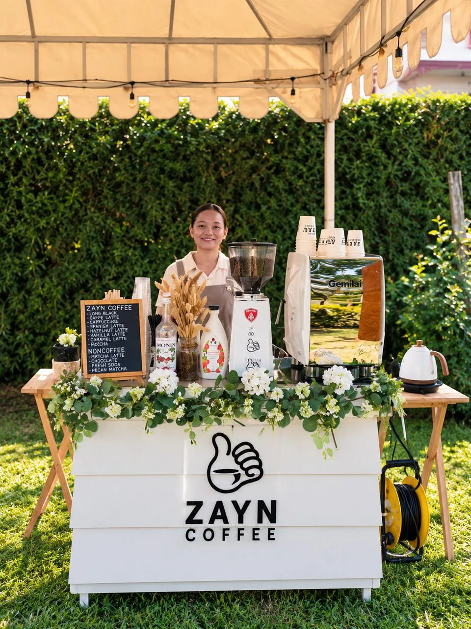 Zayn Coffee mobile coffee cart set up under a canopy on a lawn, with a barista behind the cart