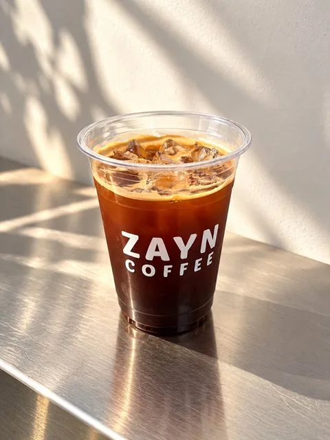 Iced Long Black in a ZAYN COFFEE cup on a steel counter in sunlight