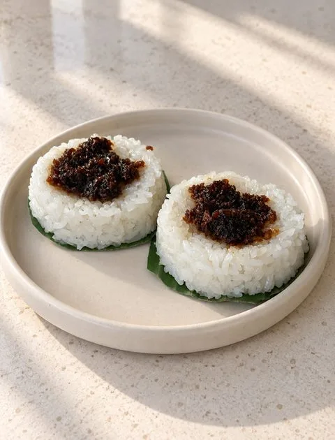 Two Puto Maya topped with latik on banana leaves on a cream plate