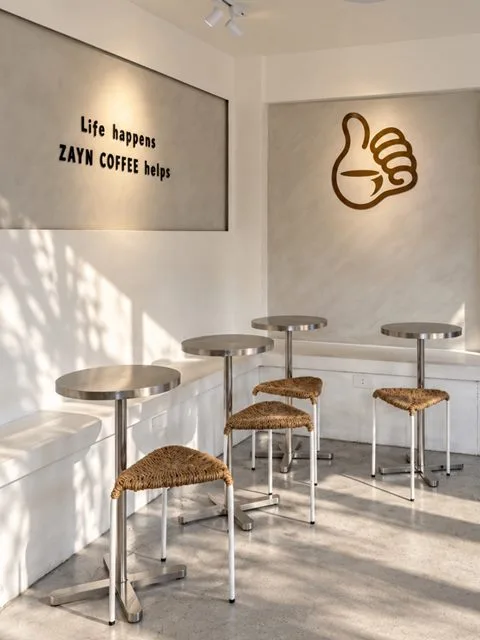 Round metal tables and woven stools in sunlight beneath a “Life happens, Zayn Coffee helps” wall sign